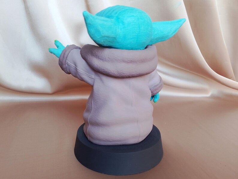 K&ouml;nnte beinhalten: Eine 3D-gedruckte Figur eines Charakters aus dem Star Wars-Franchise, bekannt als "The Child" oder "Baby Yoda". Die Figur ist in hellblau bemalt und tr&auml;gt eine braune Kapuzenrobe. Die Figur steht auf einem schwarzen runden Sockel.