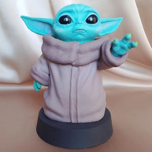 K&ouml;nnte beinhalten: Eine kleine, gr&uuml;ne und blaue Figur eines Charakters aus dem Star Wars-Franchise, bekannt als "The Child" oder "Baby Yoda". Die Figur tr&auml;gt eine braune Robe und steht auf einem schwarzen Sockel.