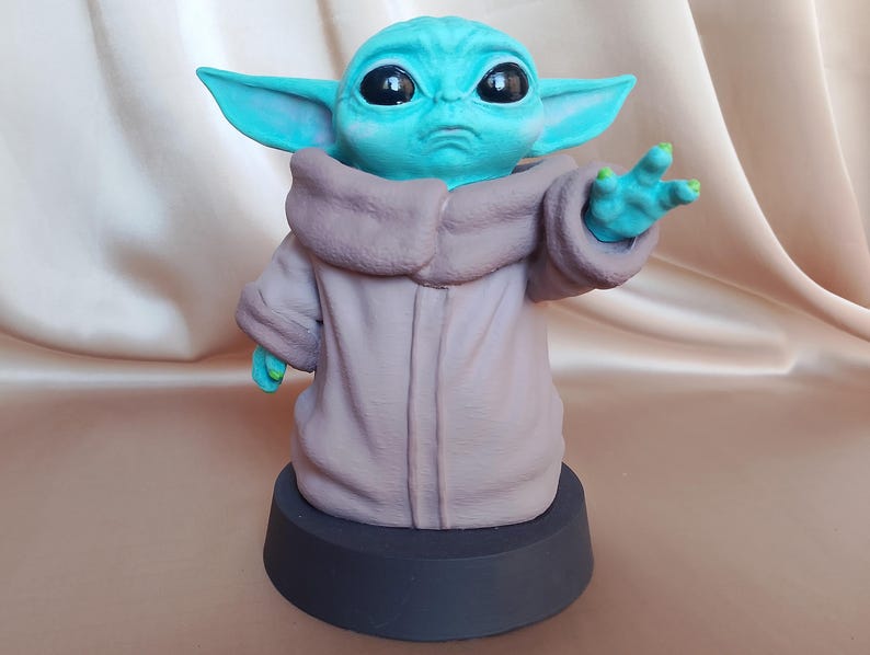 K&ouml;nnte beinhalten: Eine kleine, gr&uuml;ne und blaue Figur eines Charakters aus dem Star Wars-Franchise, bekannt als "The Child" oder "Baby Yoda". Die Figur tr&auml;gt eine braune Robe und steht auf einem schwarzen Sockel.
