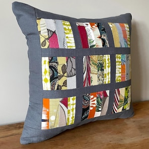 May include: Square decorative pillow with a gray border and a patchwork design. The patchwork features vertical strips of fabric in various colors and patterns, including floral and abstract designs. The pillow is resting on a wooden surface.
