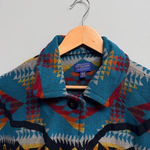 May include: A Pendleton jacket featuring a vibrant geometric pattern in teal, red, orange, yellow, and black. The jacket has a pointed collar, a single black button closure, and a blue interior label that reads Pendleton, One Size, Made in USA.