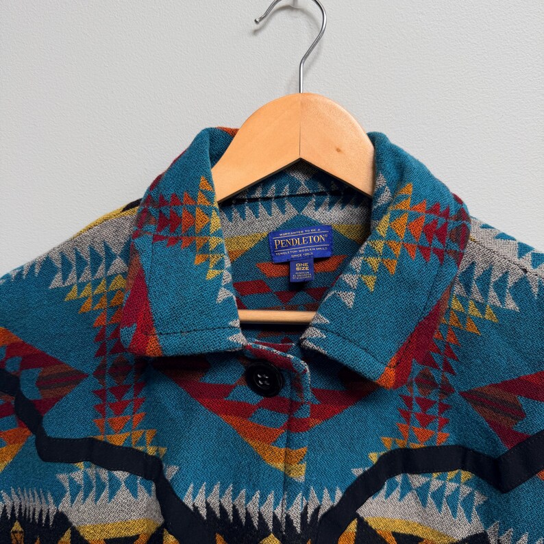May include: A Pendleton jacket featuring a vibrant geometric pattern in teal, red, orange, yellow, and black. The jacket has a pointed collar, a single black button closure, and a blue interior label that reads Pendleton, One Size, Made in USA.