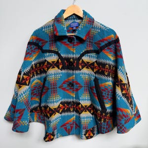 May include: A teal wool cape featuring a vibrant geometric pattern in shades of red, orange, yellow, black, and white. The garment includes a fold-over collar, four black buttons down the front, and two black-trimmed diagonal slit pockets.