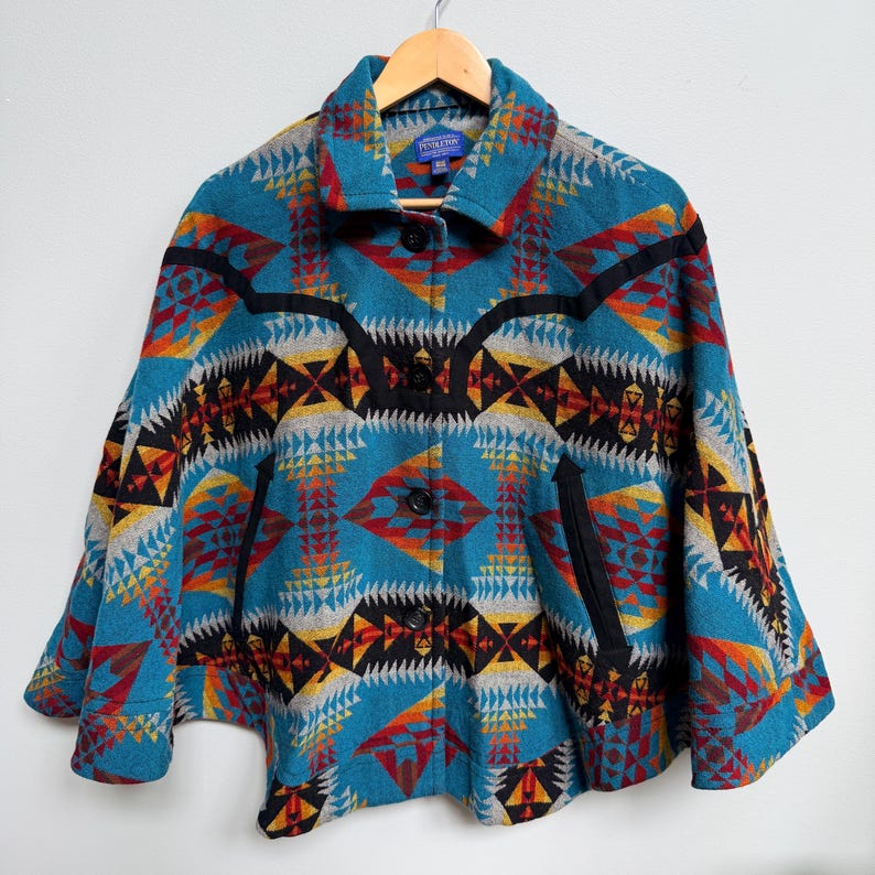 May include: A teal wool cape featuring a vibrant geometric pattern in shades of red, orange, yellow, black, and white. The garment includes a fold-over collar, four black buttons down the front, and two black-trimmed diagonal slit pockets.