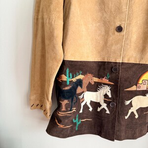 May include: A tan suede jacket with a brown lower panel featuring embroidered horses, cacti, and desert scenery. The jacket has a button-up front and long sleeves. The color palette includes tan, brown, black, white, and green.
