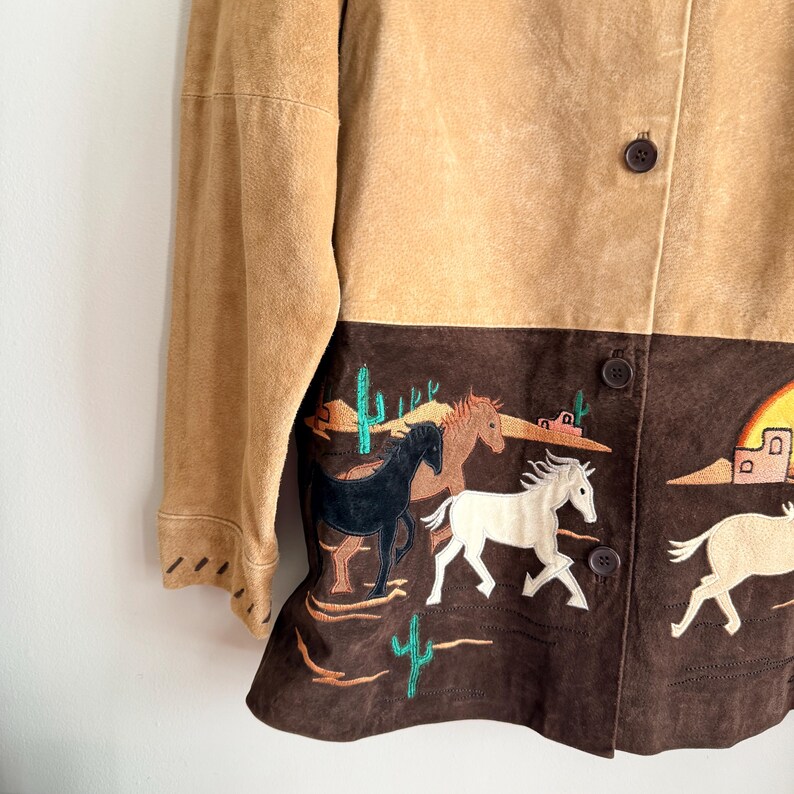 May include: A tan suede jacket with a brown lower panel featuring embroidered horses, cacti, and desert scenery. The jacket has a button-up front and long sleeves. The color palette includes tan, brown, black, white, and green.