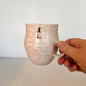 May include: A ceramic mug with a pink floral pattern and a small brown letter "L" on the side. The mug has a white handle and a rounded shape. The mug is held by a person.
