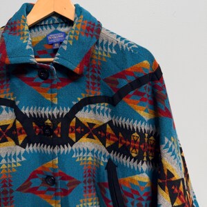 May include: A teal jacket featuring a geometric pattern in red, yellow, grey, and black. The jacket has a collar, black trim accents, and black buttons down the front. A blue Pendleton brand label is visible on the inside of the collar.