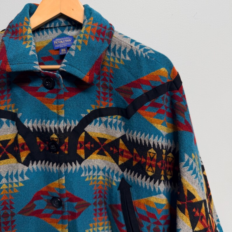 May include: A teal jacket featuring a geometric pattern in red, yellow, grey, and black. The jacket has a collar, black trim accents, and black buttons down the front. A blue Pendleton brand label is visible on the inside of the collar.