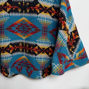 May include: A long-sleeve pullover top featuring a vibrant geometric pattern in shades of teal, black, red, yellow, and grey. The design consists of repeating diamond and triangular shapes across the fabric, with a solid black neckline trim.