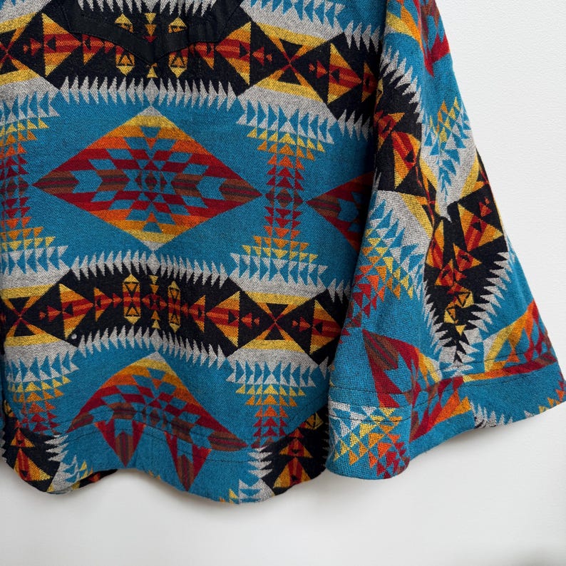 May include: A long-sleeve pullover top featuring a vibrant geometric pattern in shades of teal, black, red, yellow, and grey. The design consists of repeating diamond and triangular shapes across the fabric, with a solid black neckline trim.