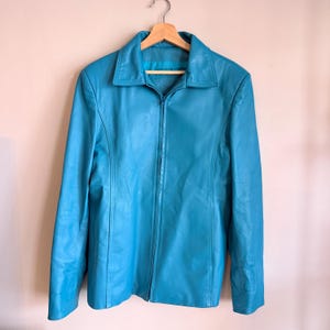 May include: A turquoise leather jacket with a collared neckline and a full-length zip. The jacket is hanging on a wooden hanger. The jacket has long sleeves and a tailored fit. The background is a neutral colour.