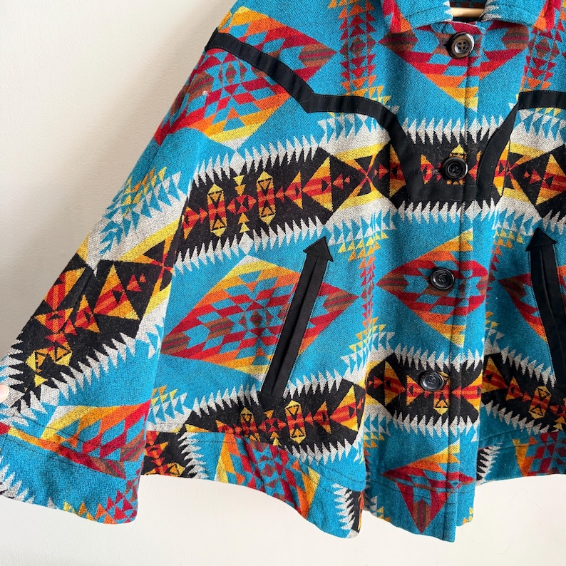 May include: A turquoise jacket featuring a vibrant geometric pattern in red, orange, yellow, black, and white. The garment includes a collar, a front button closure with dark buttons, and two slanted side pockets accented with black trim.