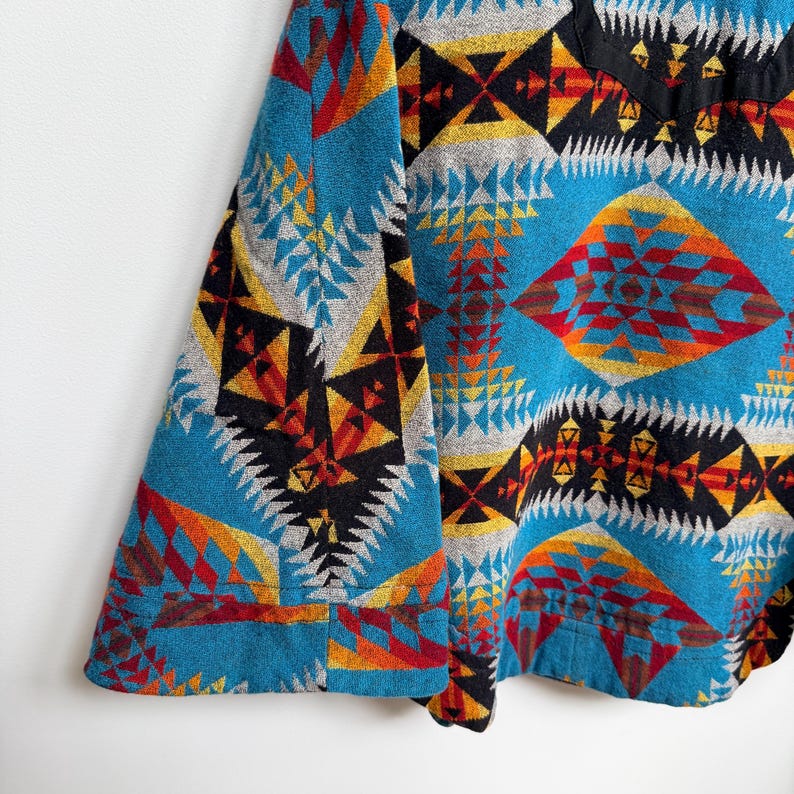 May include: A long-sleeved shirt featuring a vibrant geometric pattern in shades of turquoise, black, yellow, orange, and red. The design consists of repeating diamond and triangular shapes across the fabric, with a solid black collar detail.