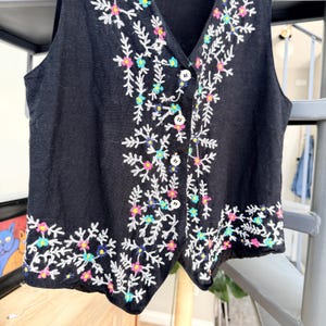May include: Black vest with a V-neck and white buttons. The vest is embroidered with a floral design in white, pink, blue, and yellow. The embroidery is along the edges and down the front.