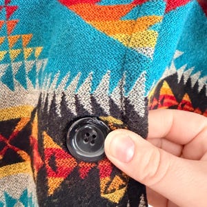 May include: A woven fabric garment featuring a vibrant geometric pattern in shades of turquoise, red, yellow, black, and white. The textile has a textured surface and includes a single, round, black four-hole button secured to the front panel.