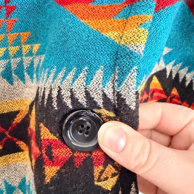 May include: A woven fabric garment featuring a vibrant geometric pattern in shades of turquoise, red, yellow, black, and white. The textile has a textured surface and includes a single, round, black four-hole button secured to the front panel.