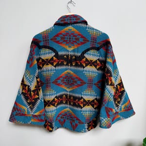 May include: A teal jacket featuring a vibrant geometric pattern in shades of orange, red, yellow, black, and grey. The garment has a wide collar, long sleeves with cuffs, and a structured back yoke, displayed hanging on a metal hanger.