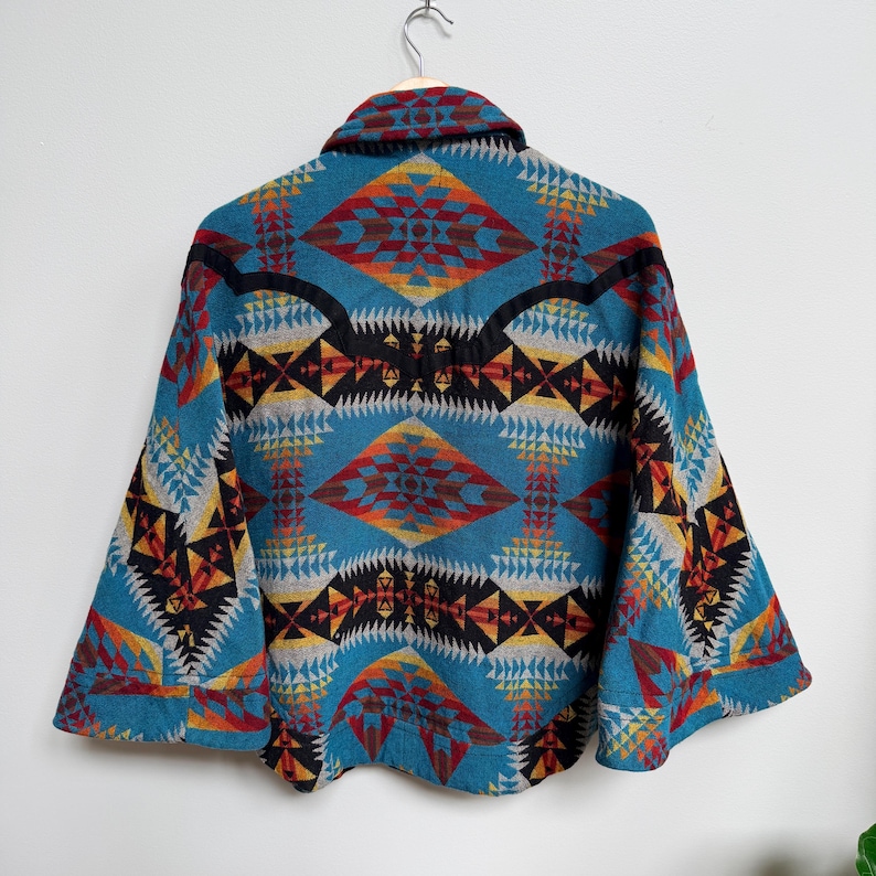 May include: A teal jacket featuring a vibrant geometric pattern in shades of orange, red, yellow, black, and grey. The garment has a wide collar, long sleeves with cuffs, and a structured back yoke, displayed hanging on a metal hanger.