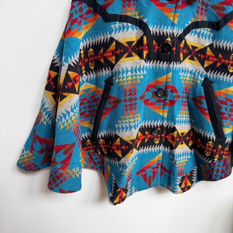 May include: A teal jacket featuring a vibrant geometric pattern in shades of red, orange, yellow, black, and white. The garment includes a black yoke, black trim along the pockets, and three black buttons down the front center closure.
