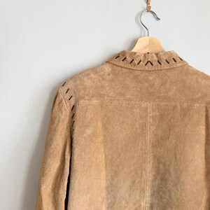 May include: A tan suede jacket hangs on a wooden hanger. The back of the jacket is visible, showcasing a stitched design along the collar and shoulders. The jacket has a classic, tailored silhouette.