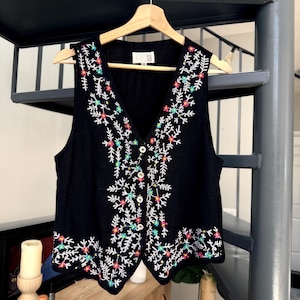 May include: Black vest with a V-neck and floral embroidery in white, pink, green, and yellow. The vest has a button-up front and is hanging on a wooden hanger.