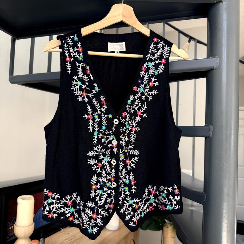 May include: Black vest with a V-neck and floral embroidery in white, pink, green, and yellow. The vest has a button-up front and is hanging on a wooden hanger.