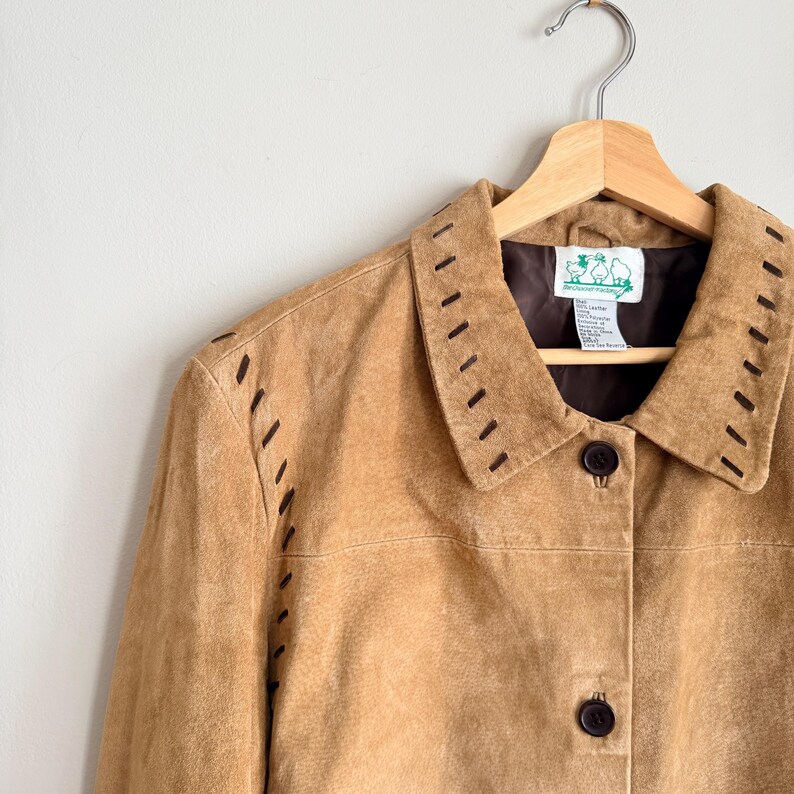 May include: A tan suede jacket with a collared neckline and button closure. The jacket features decorative stitching along the collar and seams. It hangs on a wooden hanger against a white wall.
