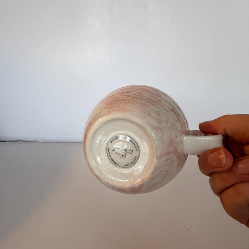 May include: A ceramic mug with a white handle and a pink and white swirled pattern. The bottom of the mug has a circular logo with a bird illustration and text. The mug is held by a hand.