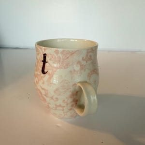 May include: Cream-colored ceramic mug with a pink floral pattern and a dark brown letter "t". The mug has a rounded shape and a curved handle. The design is a vintage style, suitable for tea or coffee.