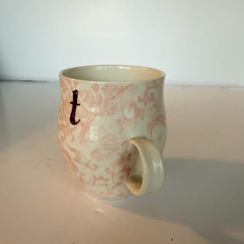 May include: Cream-colored ceramic mug with a pink floral pattern and a dark brown letter "t". The mug has a rounded shape and a curved handle. The design is a vintage style, suitable for tea or coffee.