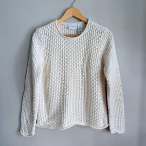 May include: Cream-coloured, textured knit jumper with a crew neck and long sleeves. The jumper is hanging on a wooden hanger against a light grey background. The knit pattern features a repeating diamond design.