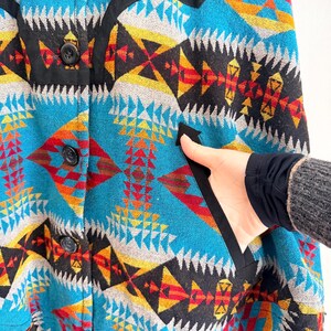 May include: A jacket featuring a vibrant geometric pattern in shades of turquoise, yellow, red, and black. The garment includes a front button closure with three black buttons and a side pocket trimmed with black fabric along the opening edge.