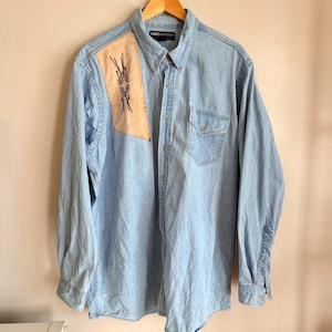May include: Pale blue denim shirt with long sleeves, a button-down collar, and two chest pockets. The shirt has a tan patch on the left shoulder with black embroidered detailing. The shirt is hanging on a wooden hanger.