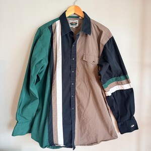 Puede incluir: Una camisa abotonada de manga larga con un diseño de bloques de color. La camisa presenta paneles verdes, negros, blancos y beige. La manga derecha tiene rayas verdes y blancas. La camisa está colgada en una percha de madera.