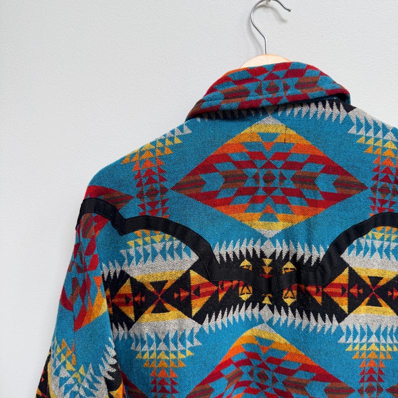 May include: A long-sleeve shirt featuring a vibrant geometric pattern in shades of turquoise, red, orange, yellow, and black. The garment includes a pointed collar and a distinct black horizontal yoke detail across the upper back and shoulders.