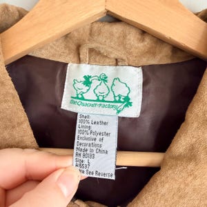 May include: Close-up of a tan leather jacket interior with a brown lining. A tag with the text "the Quacker Factory" and a duck illustration is visible. The tag also lists the shell and lining materials, size, and origin.