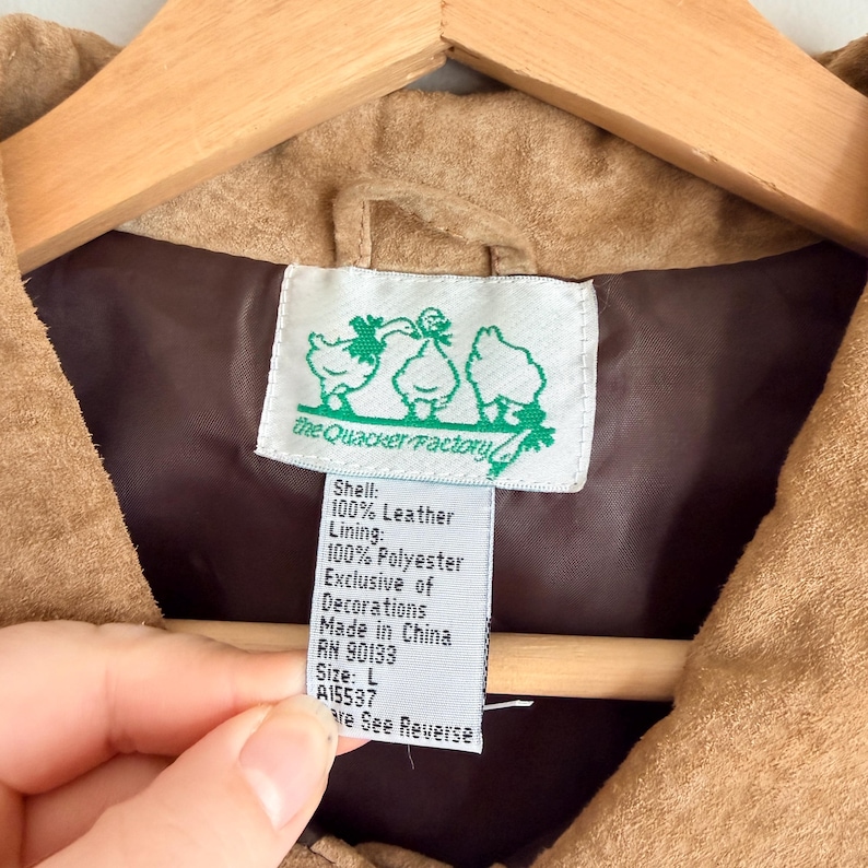 May include: Close-up of a tan leather jacket interior with a brown lining. A tag with the text "the Quacker Factory" and a duck illustration is visible. The tag also lists the shell and lining materials, size, and origin.