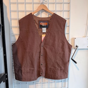 May include: A brown vest with a button-up front and two front pockets. The vest has a plaid lining and a label that reads "Cripple Creek". The vest is hanging on a wooden hanger against a white grid background.