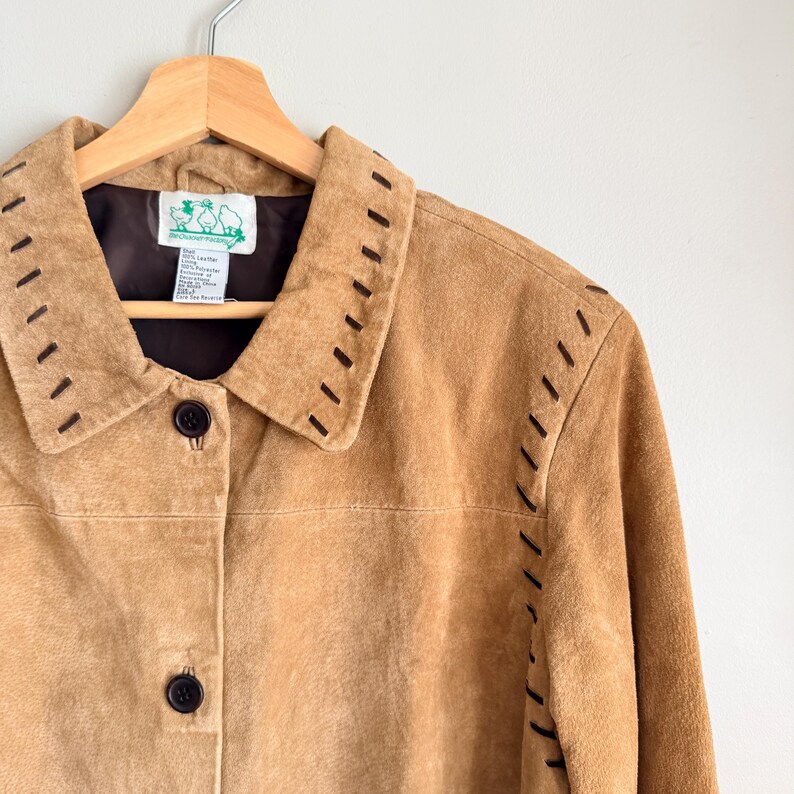 May include: A tan suede jacket with dark brown stitching details along the collar and seams. The jacket has a button-up front and is hanging on a wooden hanger. The label inside the jacket is visible.