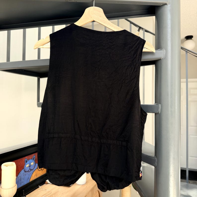 May include: Black vest with a textured fabric, hanging on a wooden hanger. The vest has a drawstring waist and a rounded bottom hem. The back of the vest is visible, showing the garment's construction and design. The vest is sleeveless.