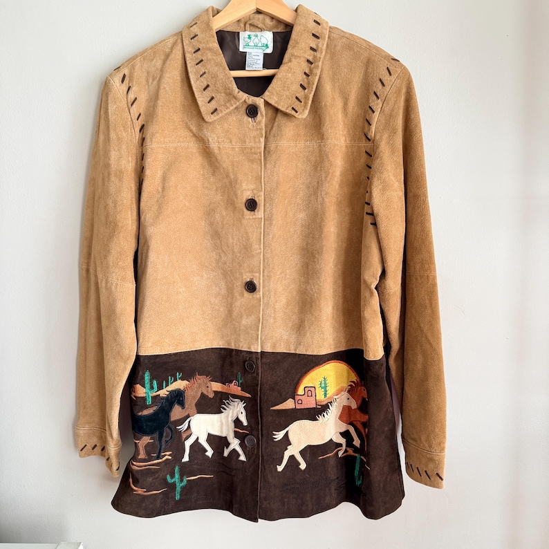 May include: A tan suede jacket with a brown bottom panel featuring embroidered horses, cacti, and a desert scene. The jacket has a collared neckline, button closures, and decorative stitching along the seams.