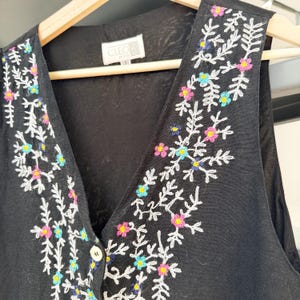May include: Black vest with white floral embroidery and colorful accents of pink, blue, and yellow. The vest has a V-neck and a button closure. The label reads "CLEO".