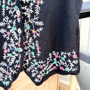 May include: Black vest with white floral embroidery. The embroidery features colorful accents in pink, blue, and yellow. The vest has a button-up front.