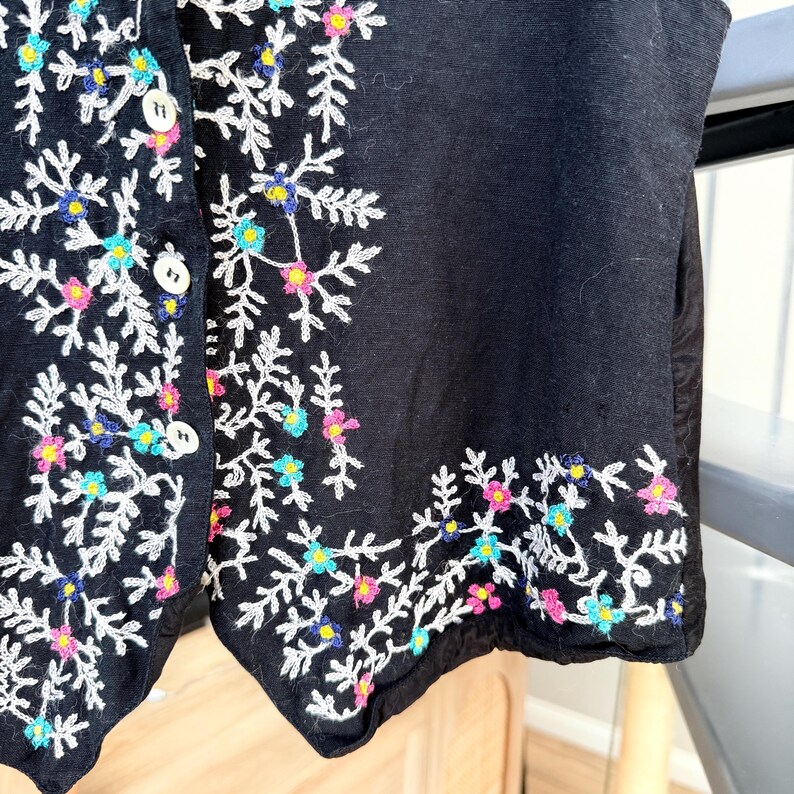 May include: Black vest with white floral embroidery. The embroidery features colorful accents in pink, blue, and yellow. The vest has a button-up front.