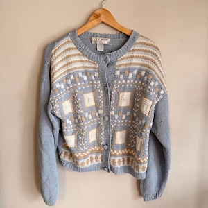May include: A light blue Studio cardigan with a round neck and button closure. The sweater features a geometric pattern in cream and tan, with ribbed cuffs and a ribbed hem. The cardigan is hanging on a wooden hanger.
