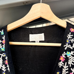 May include: Black embroidered vest with colorful floral and leaf designs. The vest is hanging on a wooden hanger. The label reads "CLEO PETITES" and size "S".