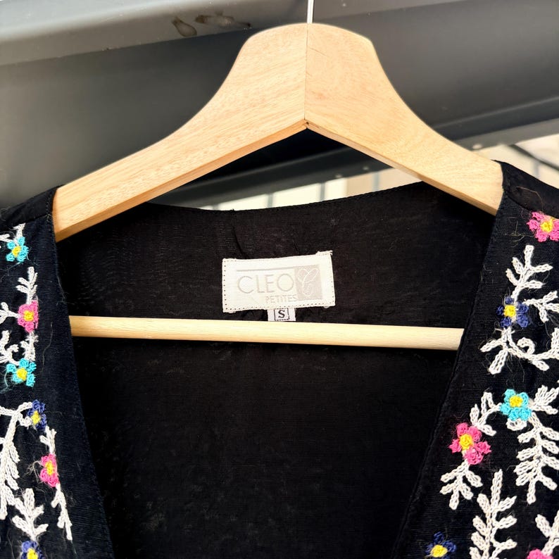May include: Black embroidered vest with colorful floral and leaf designs. The vest is hanging on a wooden hanger. The label reads "CLEO PETITES" and size "S".