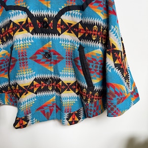 May include: A turquoise jacket featuring a vibrant geometric pattern in shades of red, yellow, black, and white. The garment includes a black yoke detail, three black buttons down the front, and two vertical side pockets with black trim.