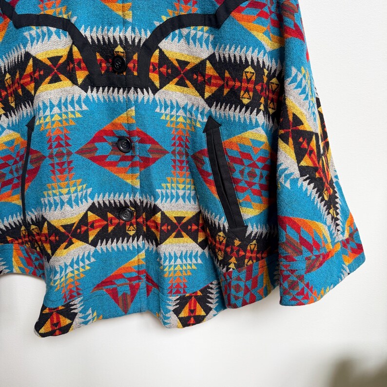 May include: A turquoise jacket featuring a vibrant geometric pattern in shades of red, yellow, black, and white. The garment includes a black yoke detail, three black buttons down the front, and two vertical side pockets with black trim.